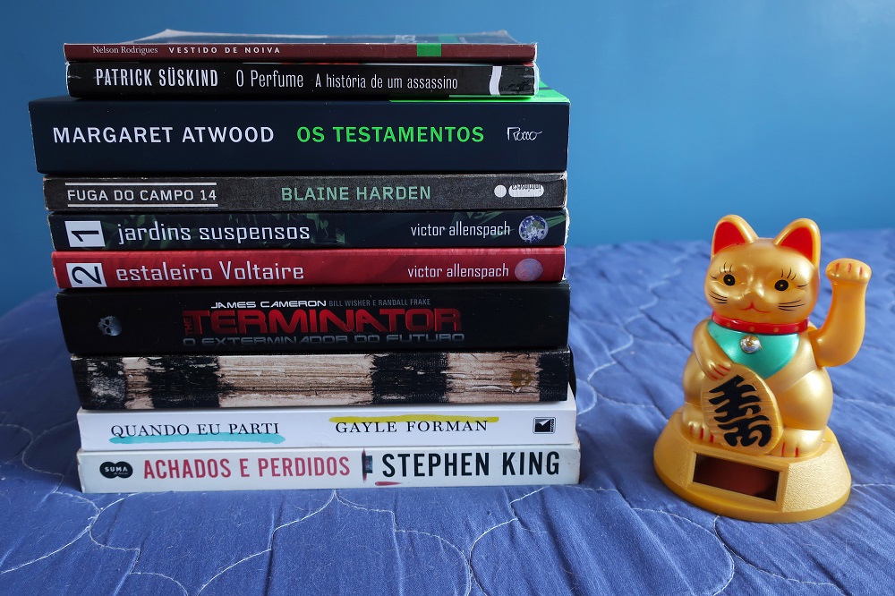 Livros empilhados sobre a cama com gato de plástico ao lado. Leituras de 2022 do blog Gato que Flutua, por Debb Cabral